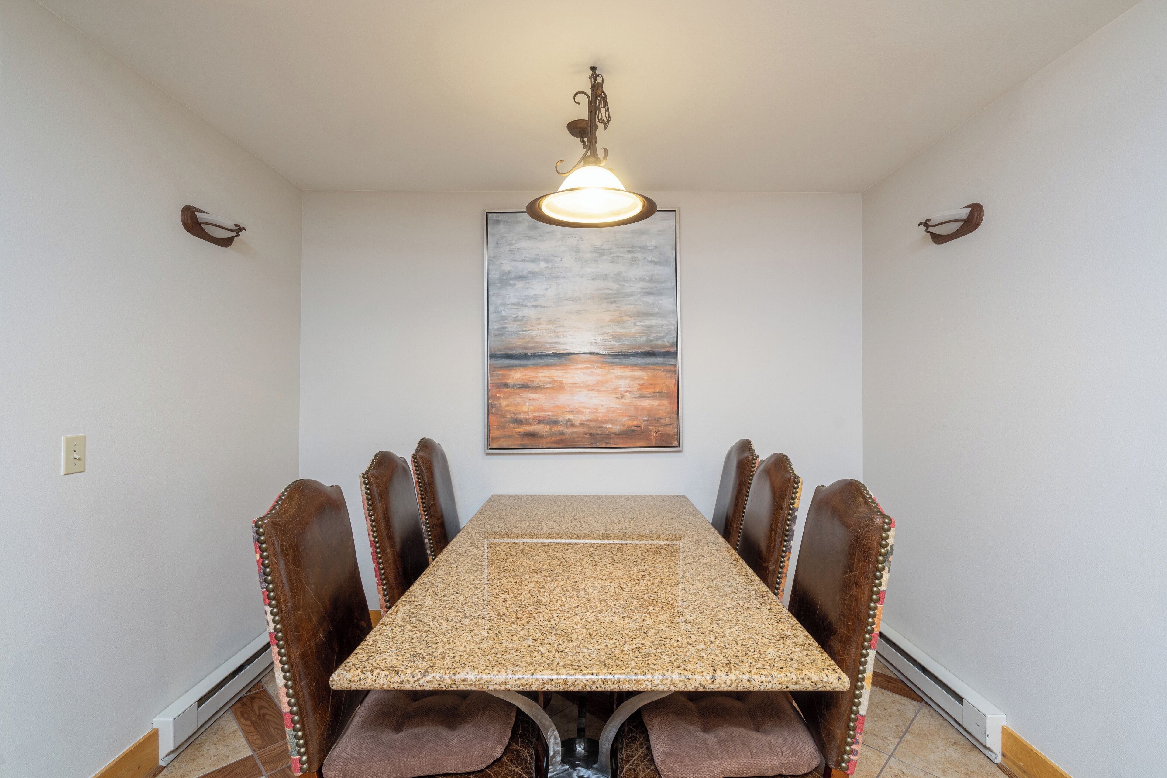 A dining room with a rectangular table, six chairs, a pendant light, and a painting of a sunset on the wall.
