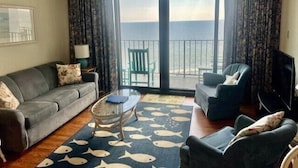 A living room with a sofa, armchairs, a glass coffee table, and a view of the sea through large sliding doors.