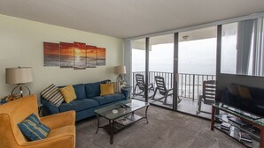 Een woonkamer met een bank, fauteuil, salontafel en tv-meubel. Drie schilderijen hangen aan de muur en er is een balkon met stoelen.