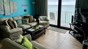 Un salon comprenant un canapé, des fauteuils, une table basse et un téléviseur. Il y a un balcon avec vue sur l’océan.