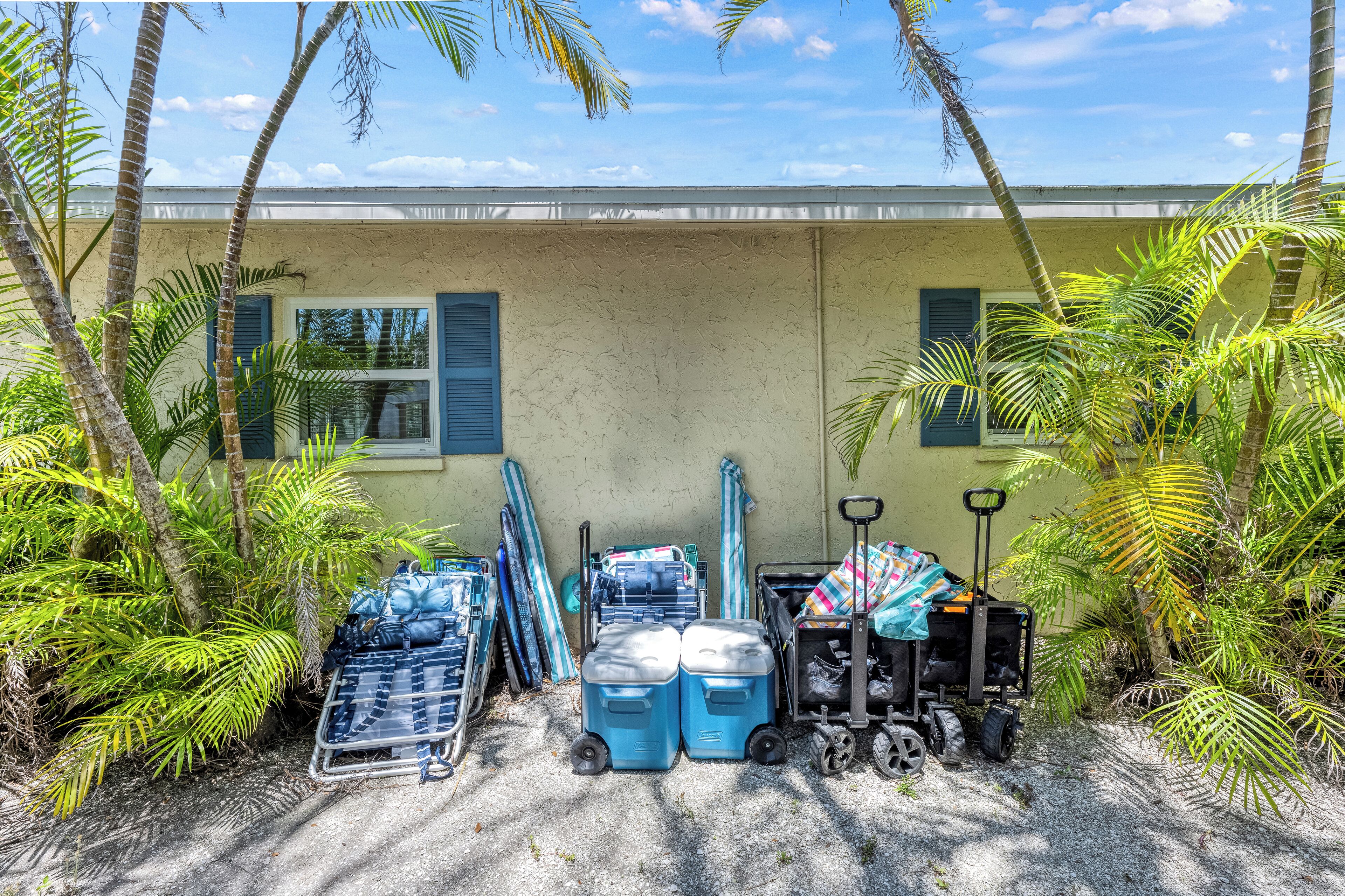 A building with a beige exterior and blue shutters, surrounded by palm trees. There are two blue coolers, a luggage cart, and a covered item on the ground.