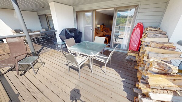 En træterrasse med et bord med glasplade og stole, en grill og et stabel med brænde.