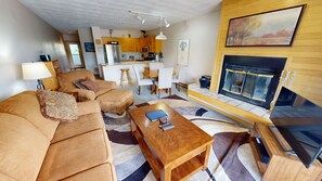 A modern living room with a sofa, coffee table, and fireplace. There is a kitchen in the background with wooden cabinets and a dining area.