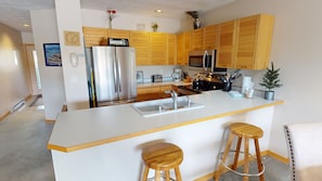 A modern kitchen with wooden cabinets, stainless steel appliances, a central island with stools, and a view of the adjacent living area.
