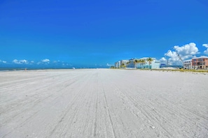 A wide sandy beach with a clear sky, a few buildings in the background, and a distant horizon.