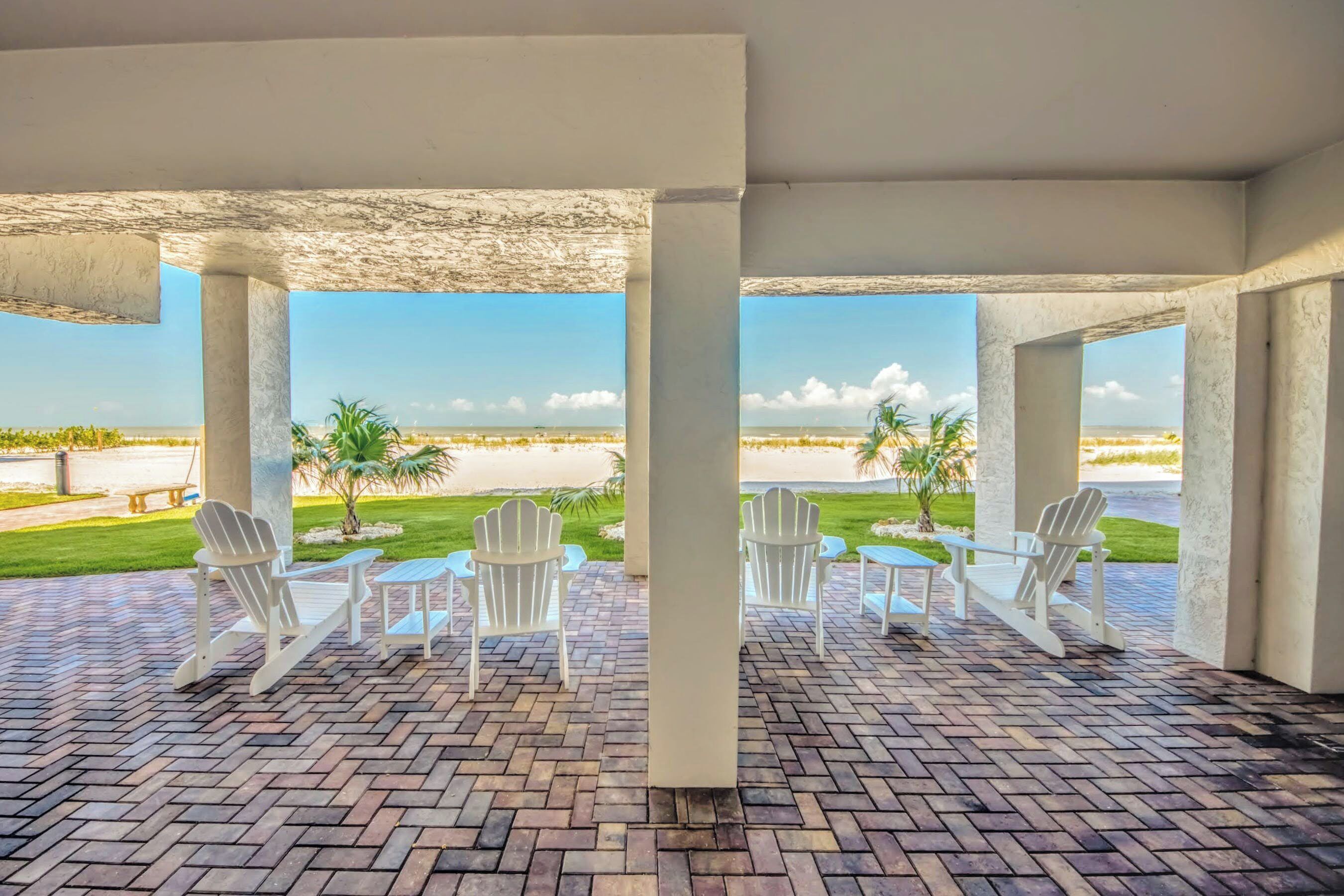 A covered outdoor area with brick flooring, white Adirondack chairs, and a view of a beach.