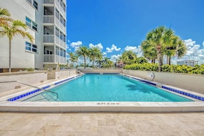A swimming pool with a clear blue water surface, surrounded by palm trees and a multi-story building with balconies. - Gateway Villas #199 Lovely Beachfront Condo (Fort Myers Beach)
