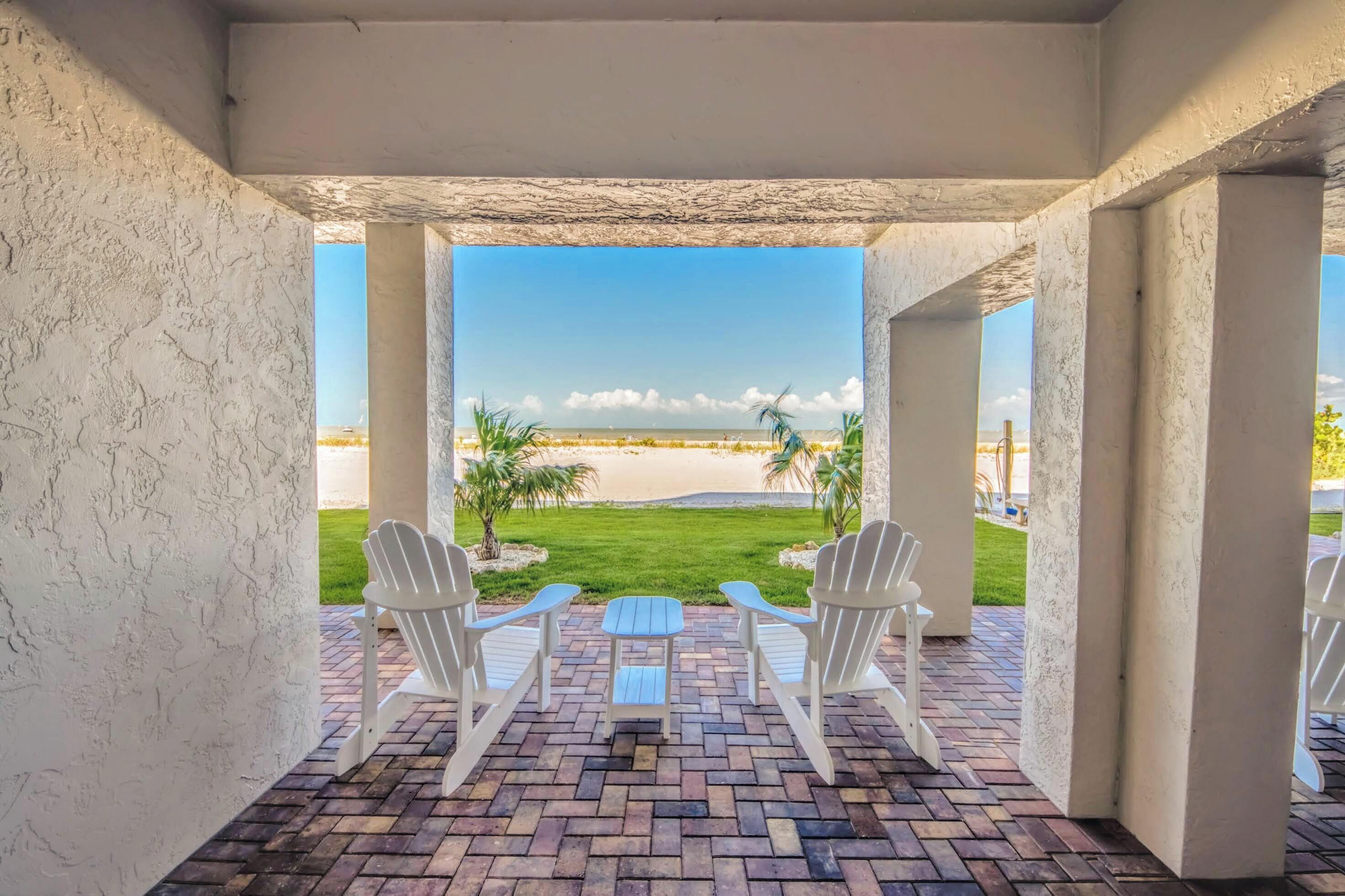 A covered patio with brick flooring, white Adirondack chairs, and a view of a beach.