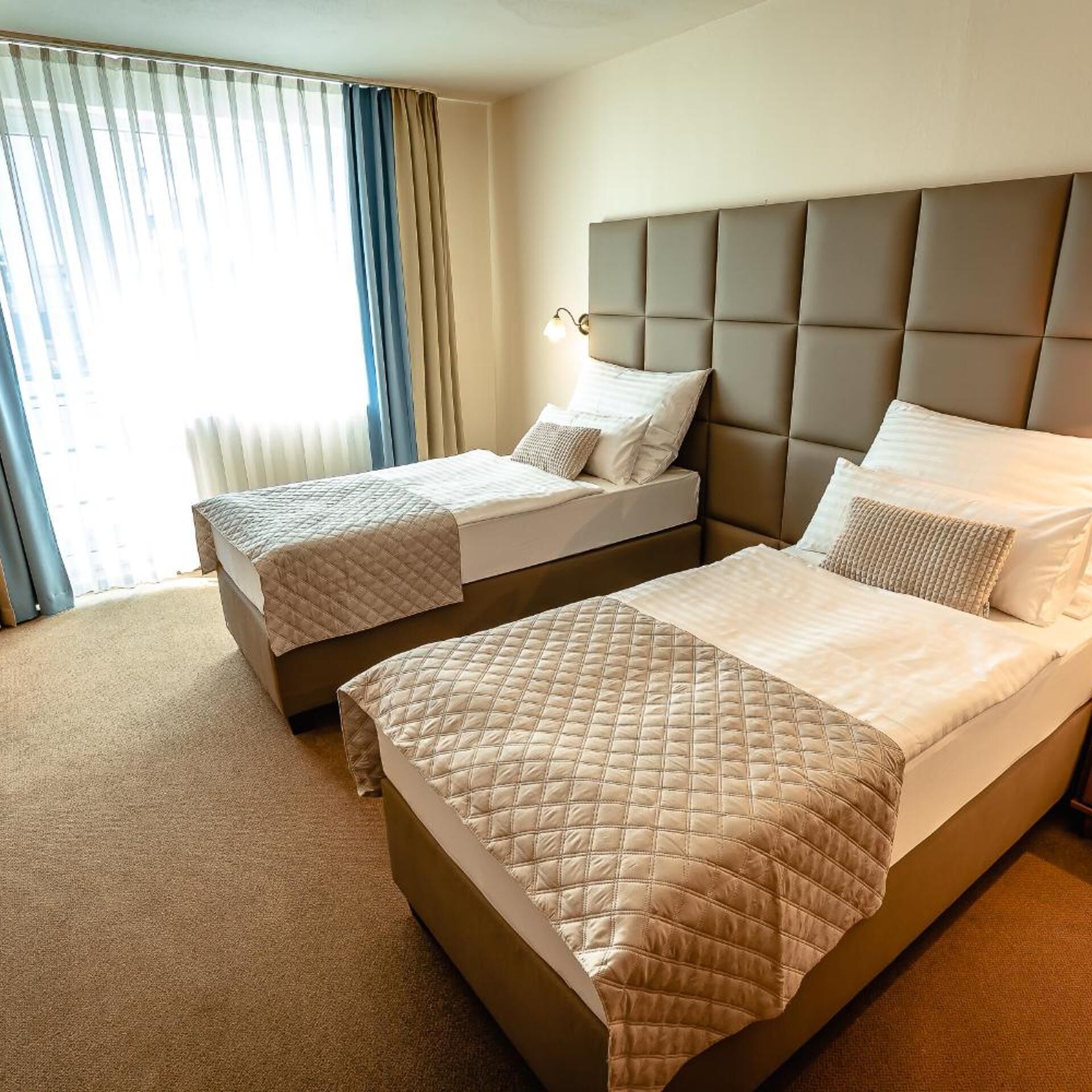 Habitación de hotel con dos camas, un ventanal grande con cortinas y un cabecero.