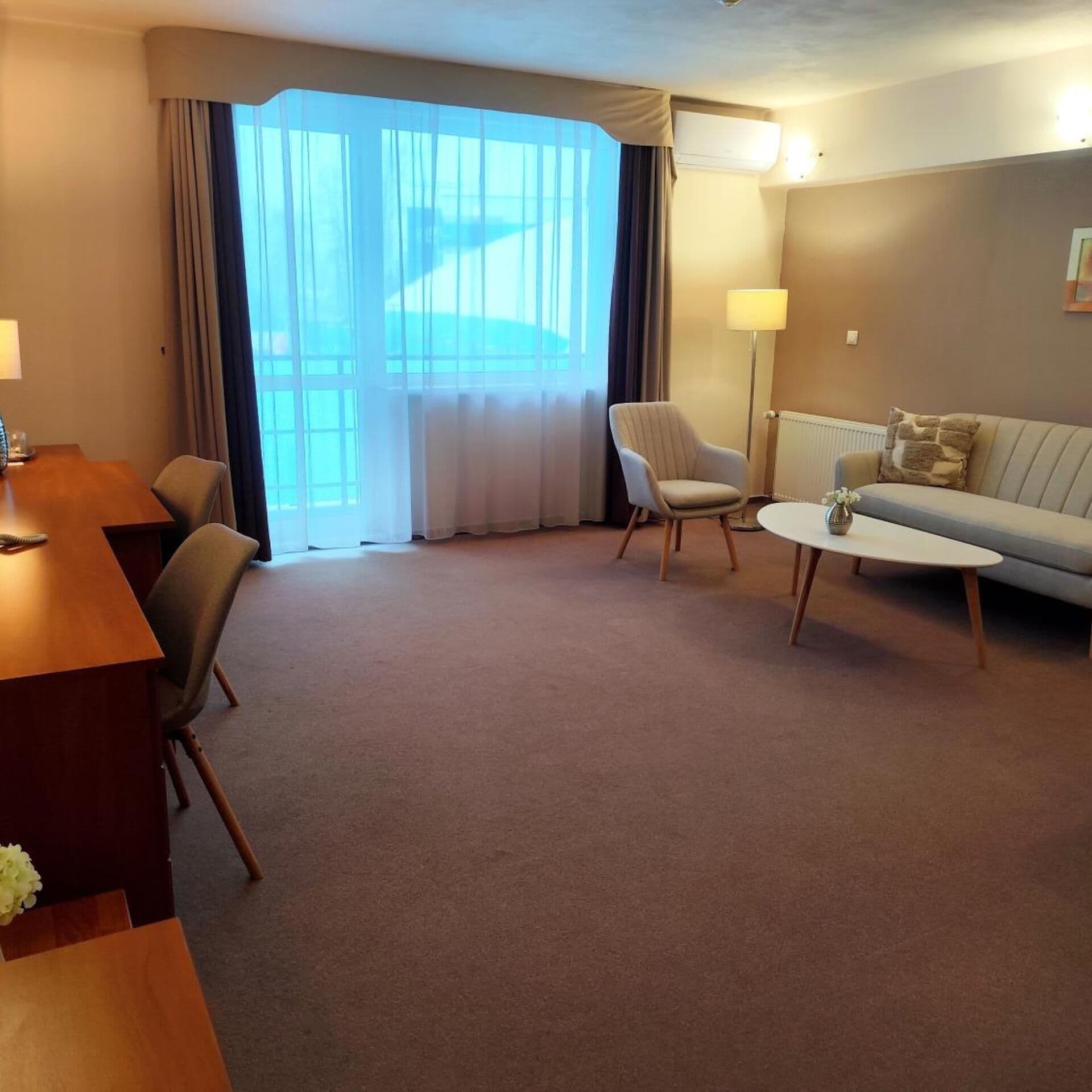Habitación de hotel con un escritorio de madera, dos sillas, un sofá, una mesita y un ventanal grande con cortinas.