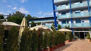 Un edificio moderno con varios pisos, mesas al aire libre con sombrillas y plantas en macetas.