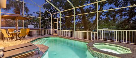 A swimming pool with a sunken hot tub, surrounded by a covered patio with chairs and an umbrella.