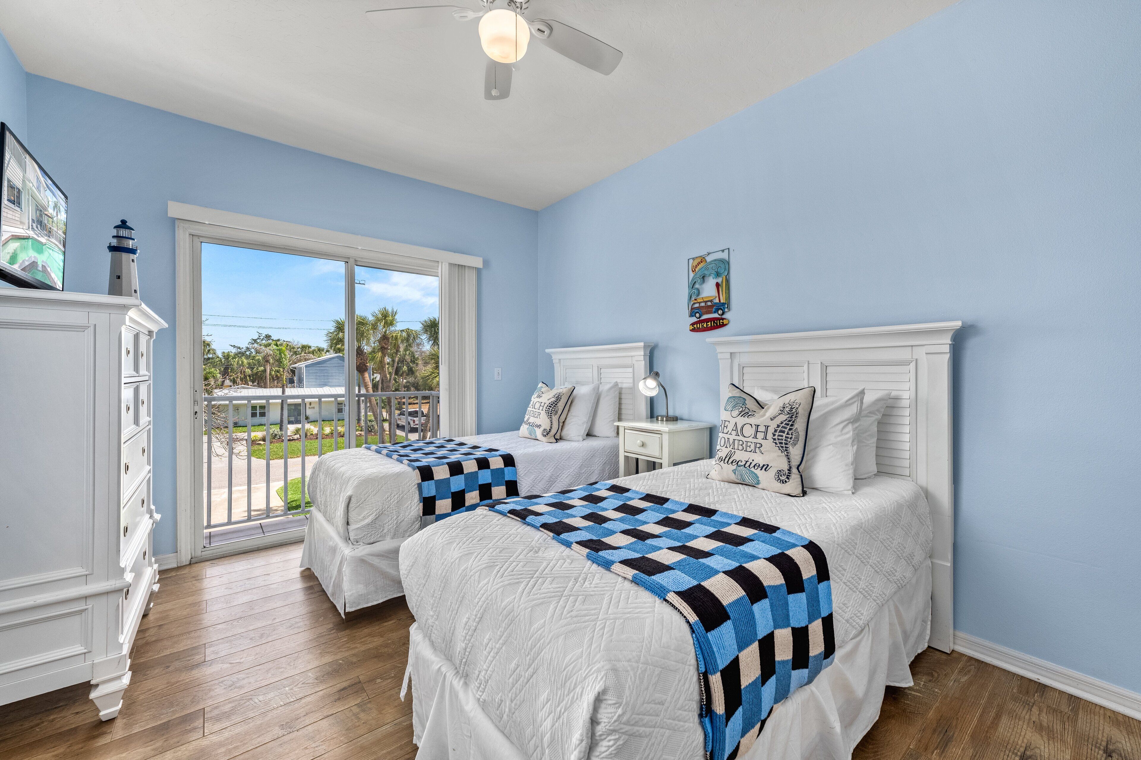 A bedroom with two beds, a window with a view of palm trees, a ceiling fan, and a dresser.