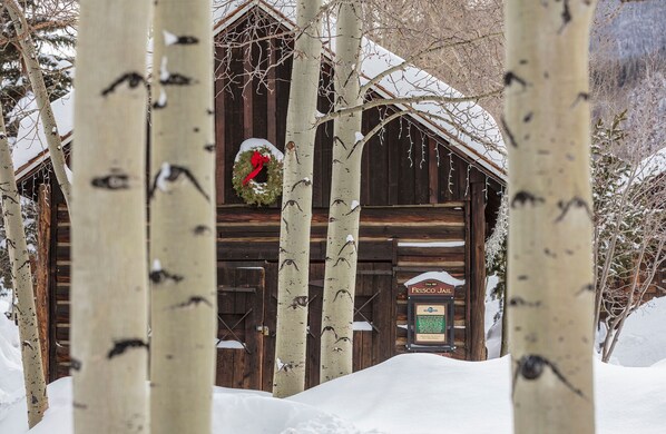 A wooden cabin with a sign reading 'Frisco Jail,' surrounded by snow-covered ground and birch trees.