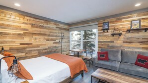 A modern room with a wooden wall, a bed with an orange blanket, a gray sofa with red cushions, and a small table. - Riverside Hotel Room With Split-King Bed (Telluride)