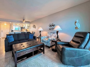 A living room with a sofa, armchair, coffee table, and a patterned armchair. There is a kitchen visible in the background.