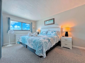 A bedroom with a bed, a bedside table with a lamp, a window with curtains, and a wall-mounted picture.