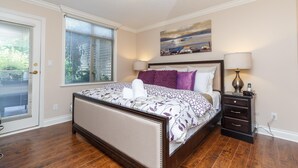 A bedroom with a bed, a nightstand, a lamp, a painting, and a window with blinds.