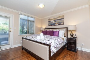 A bedroom with a bed, a nightstand, a lamp, a painting, and a window with blinds. - Versatile Garden-Level Suite w/ Natural Light (Victoria)