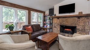 A living room with a stone fireplace, a flat-screen TV, a bookshelf, and a window with a view of trees. - Sleep Six in This Private Lofted Room With a Kitchenette (Telluride)