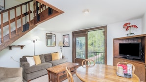A living room with a wooden staircase, a sofa, a dining table, chairs, a TV, and a window with a view of greenery. - Sleep Six in This Private Lofted Room With a Kitchenette (Telluride)