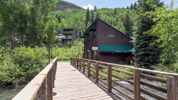 A wooden bridge leading to a lodge with a green roof, surrounded by dense forest and a river.