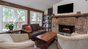 A living room with a stone fireplace, a flat-screen TV, a bookshelf, and a window with a view of trees.