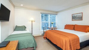A room with two beds, a desk, a chair, a lamp, and a window with a view of trees. - Mountainside Inn Room With Kitchenette (Telluride)