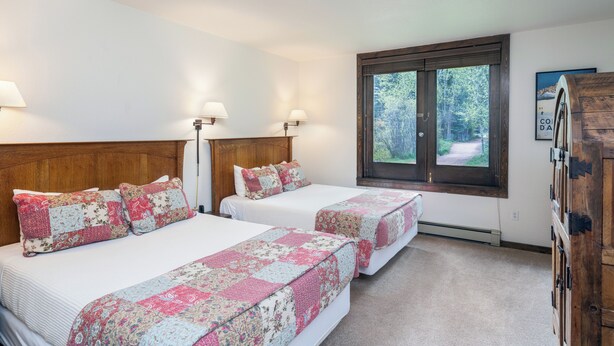 Une chambre avec deux lits, une tête de lit en bois, une grande fenêtre donnant sur les arbres et une armoire en bois.