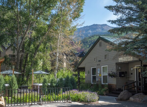A mountain lodge with a welcoming sign, surrounded by lush greenery and a view of mountains.