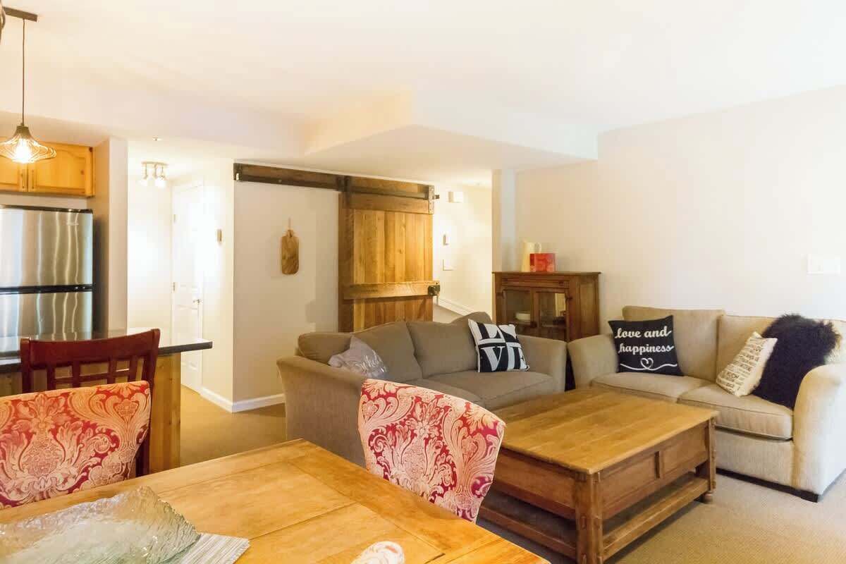 A living room with a sofa, coffee table, and chairs. There is a kitchen area with a refrigerator and cabinets.