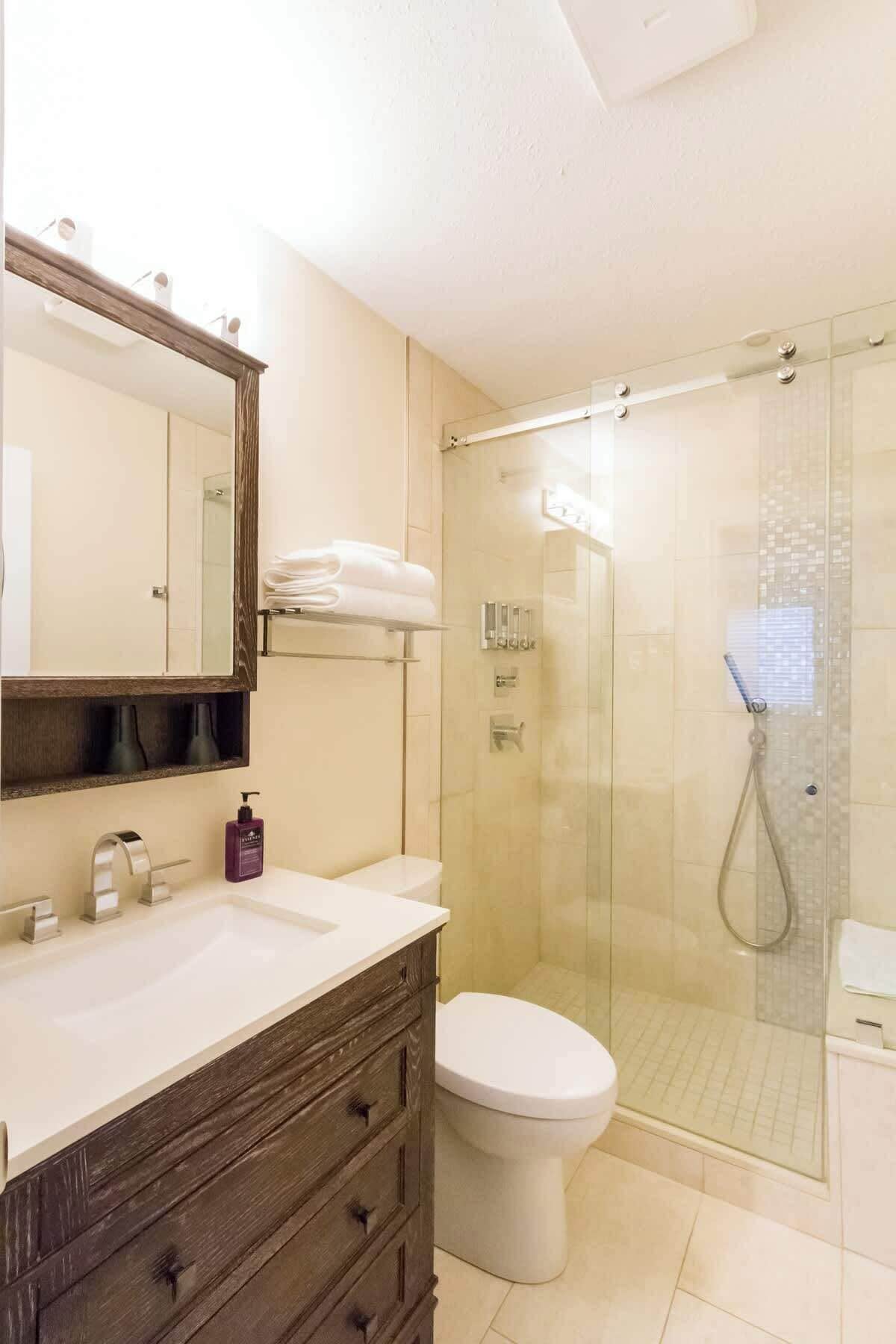 Un bagno con doccia in vetro, un WC bianco, un mobile bagno in legno con specchio e uno scaffale con asciugamani piegati.