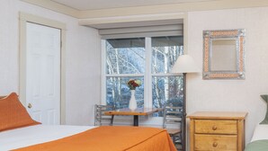 A hotel room with a bed, a desk with a chair, a window with a view of trees, a dresser, and a mirror on the wall. - Mountainside Inn Hotel Room With Kitchenette (Telluride)