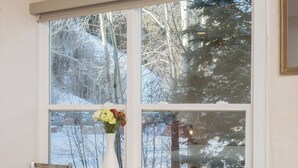 A room with a wooden table and chairs, a white vase with flowers, and a large window with a view of trees and snow. - Mountainside Inn Hotel Room With Kitchenette (Telluride)
