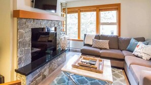 A living room with a stone fireplace, a sofa, a coffee table, and a TV mounted above the fireplace. - Ski Home, Free Shuttle, Communal Hot Tub (Whistler)