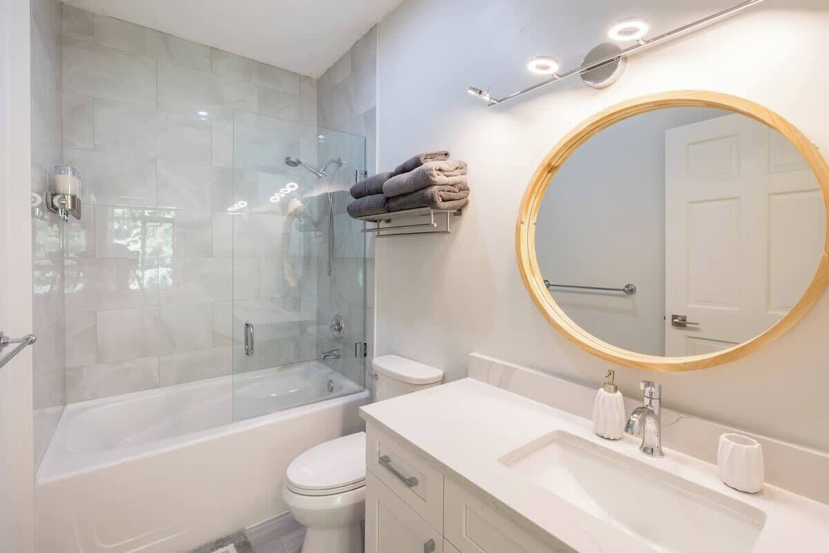 A modern bathroom with a glass-enclosed shower, a white toilet, a vanity with a sink, and a round mirror.