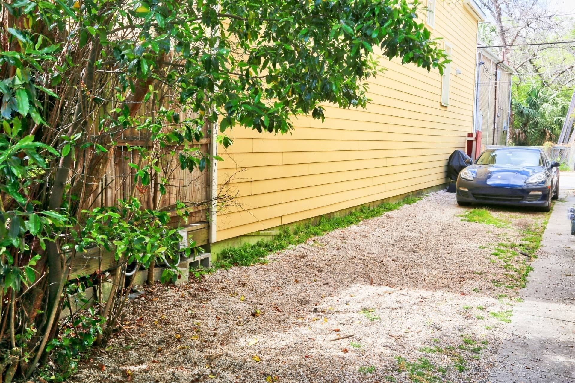 A gravel driveway leading to a parked car, a yellow house, and a wooden fence with greenery.