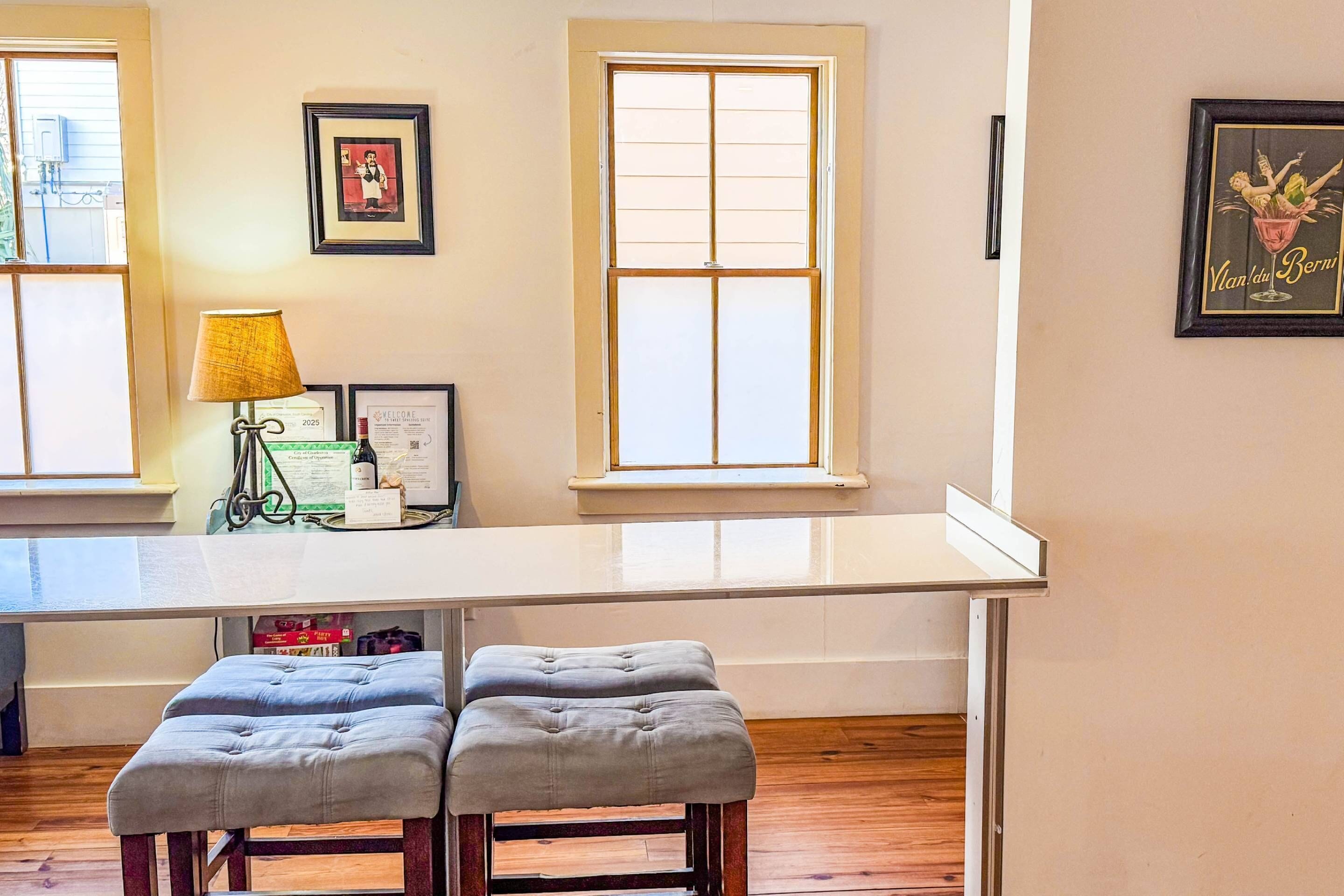 A kitchen with a white countertop, two grey cushioned stools, a table lamp, framed artwork, and a window with a view.