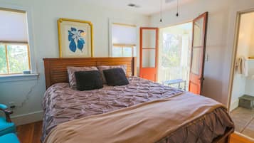 Une chambre avec un lit en bois, deux oreillers, une fenêtre avec des stores, un tableau encadré au mur et une porte donnant sur l’extérieur.