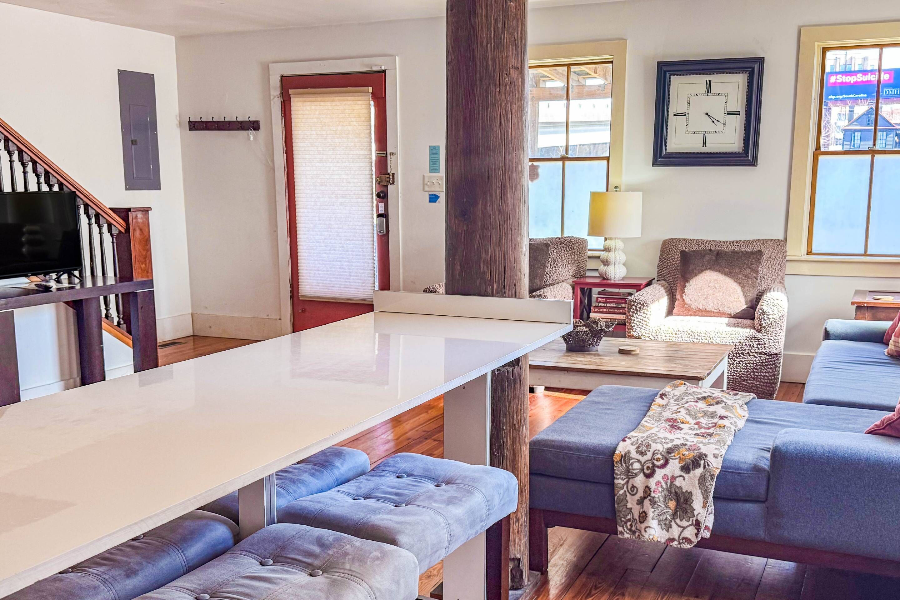 Un salon moderne avec une grande table blanche, des canapés bleus, un escalier en bois et un téléviseur.