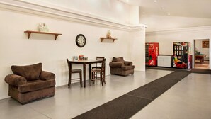Un vestíbulo amplio con techo alto, una mesa central con sillas y dos sofás. Hay relojes y estanterías con objetos decorativos en las paredes. Se ve una máquina expendedora de Coca-Cola al fondo.