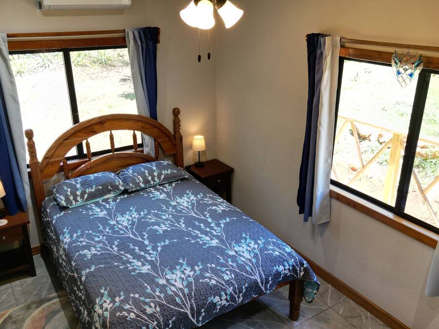 A bedroom with a bed, two bedside tables with lamps, a window with curtains, and a view of the outdoors.