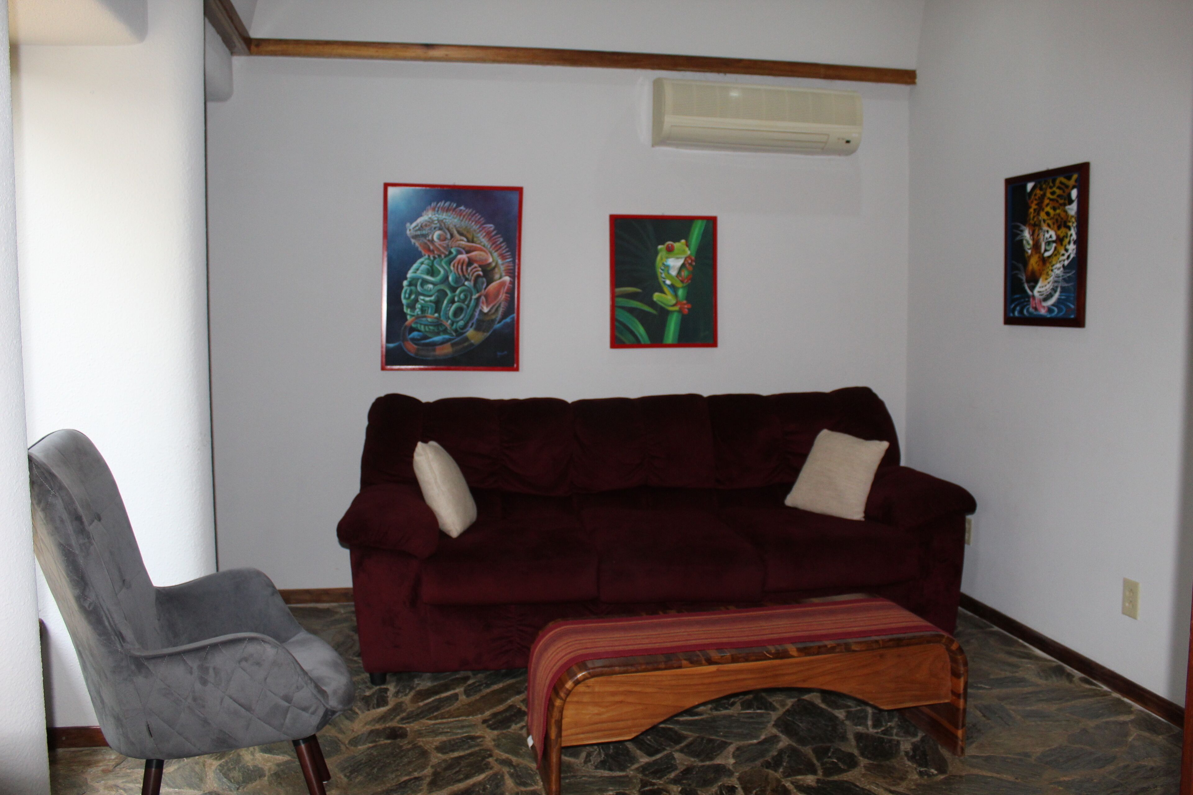 A living room with a dark red sofa, a grey armchair, and a wooden bench. There are three framed artworks on the wall.