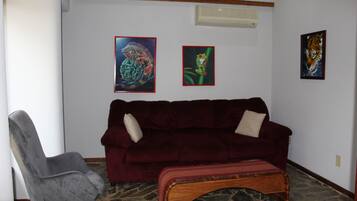 A living room with a dark red sofa, a grey armchair, and a wooden bench. There are three framed artworks on the wall.
