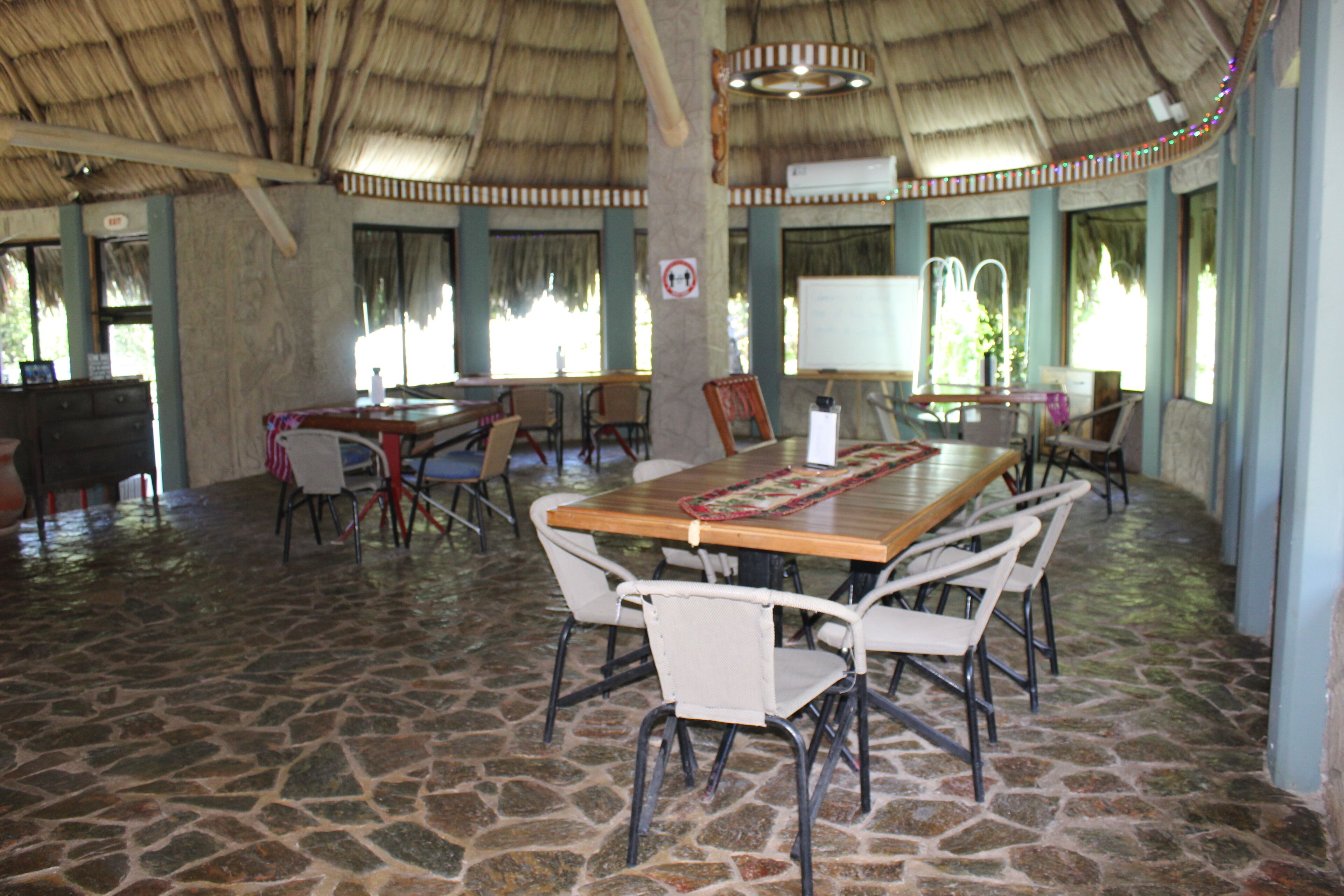 Un espace intérieur vaste, doté d’un sol en pierre, d’une table en bois entourée de chaises et d’un toit de chaume.