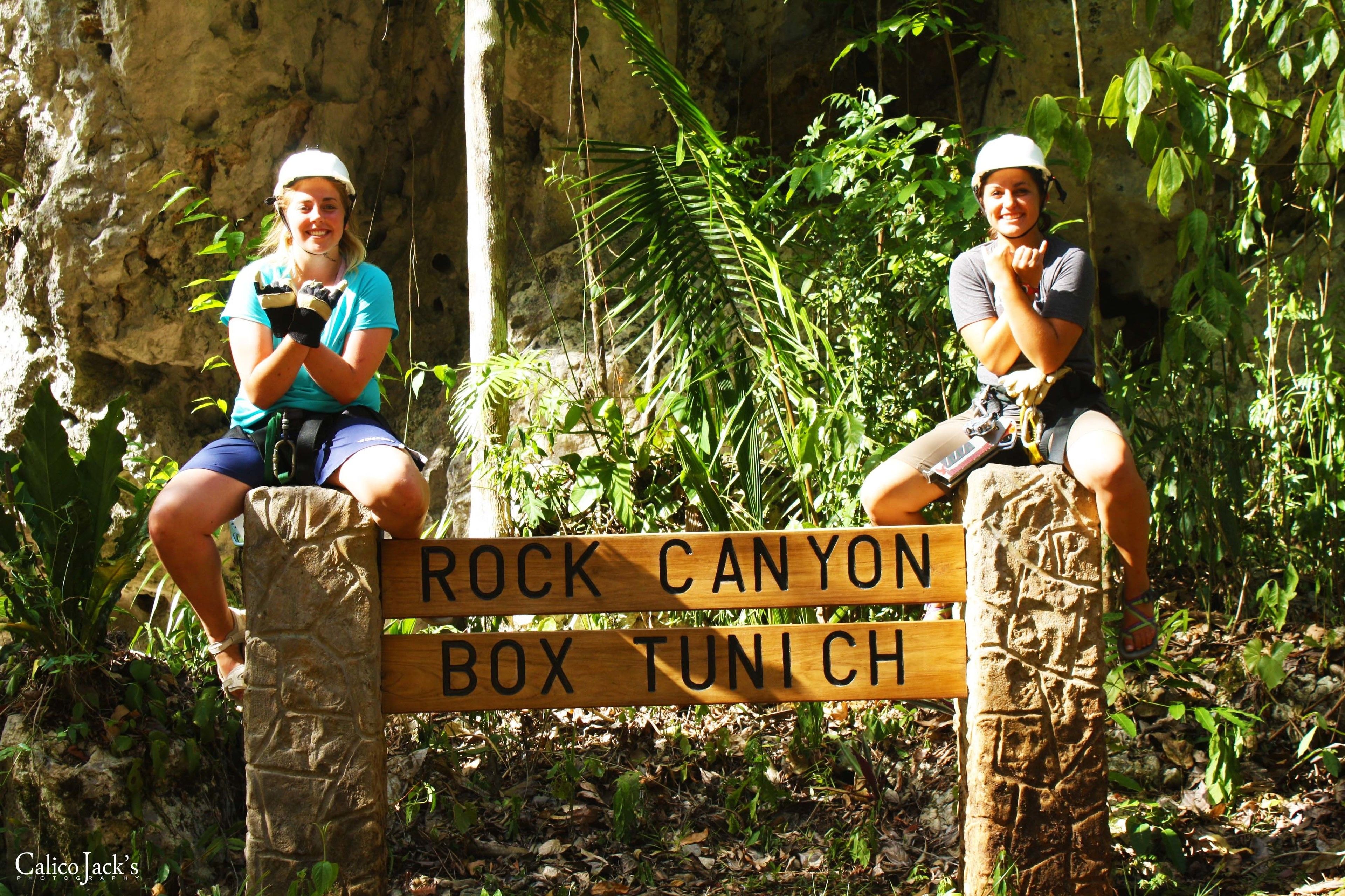 Two people sitting on a wooden bench with a sign that reads "ROCK CANYON BOX TUNICH".