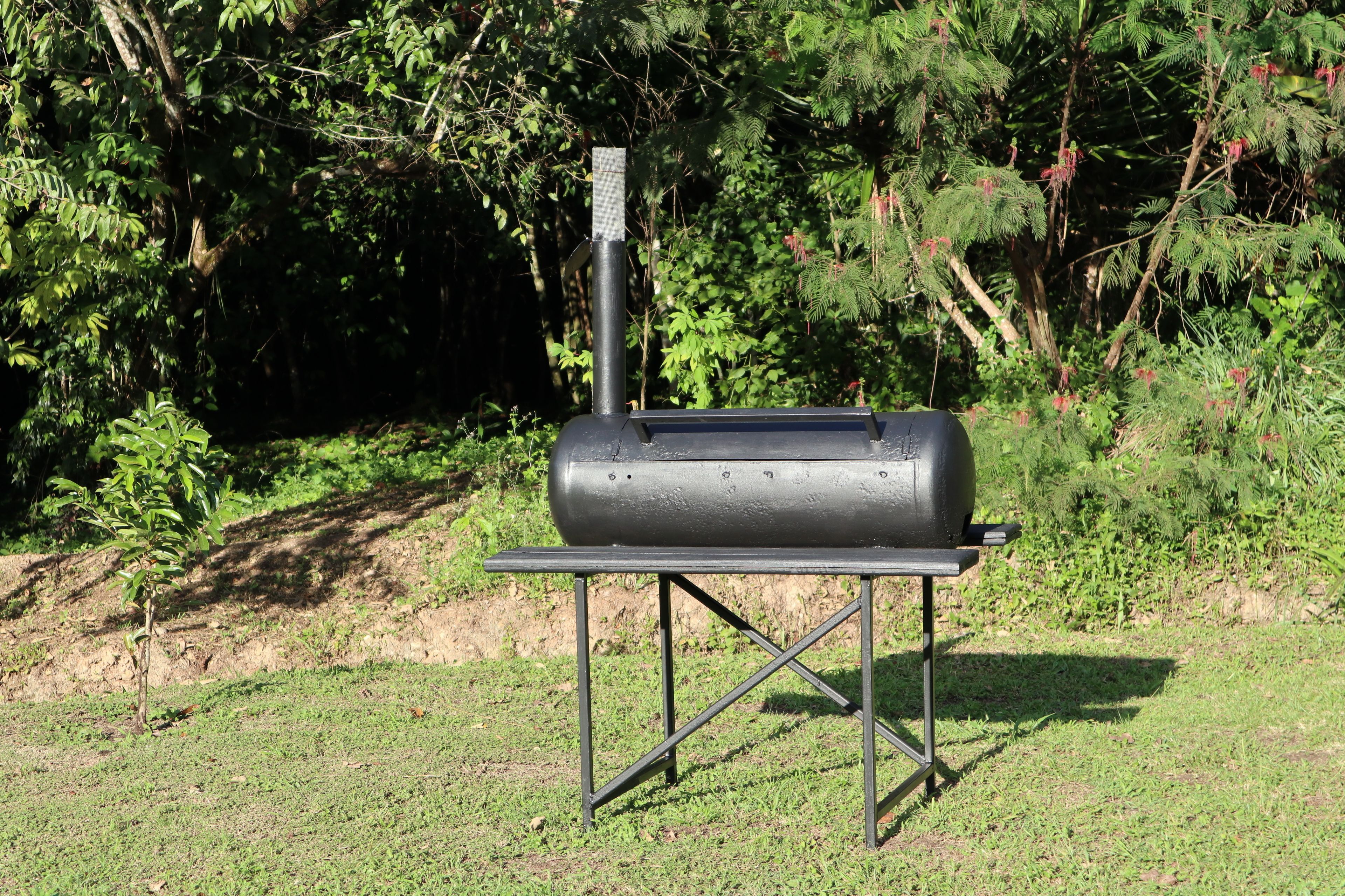 Un barbecue portable, posé sur un support métallique, se trouve dans une zone herbeuse, avec un feuillage dense en arrière-plan.