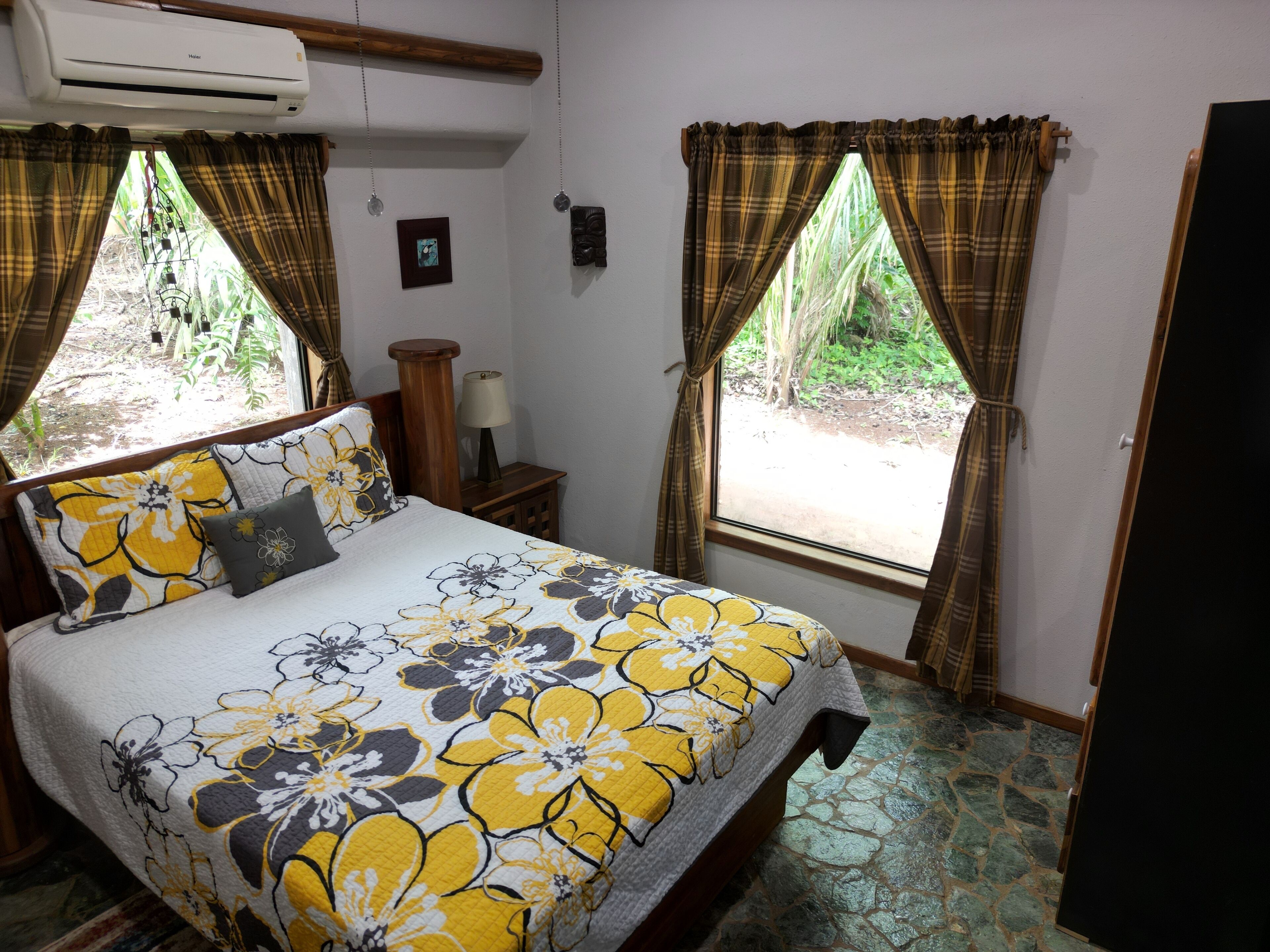 A bedroom with a bed, two windows with curtains, a bedside table with a lamp, and a wall-mounted air conditioner.