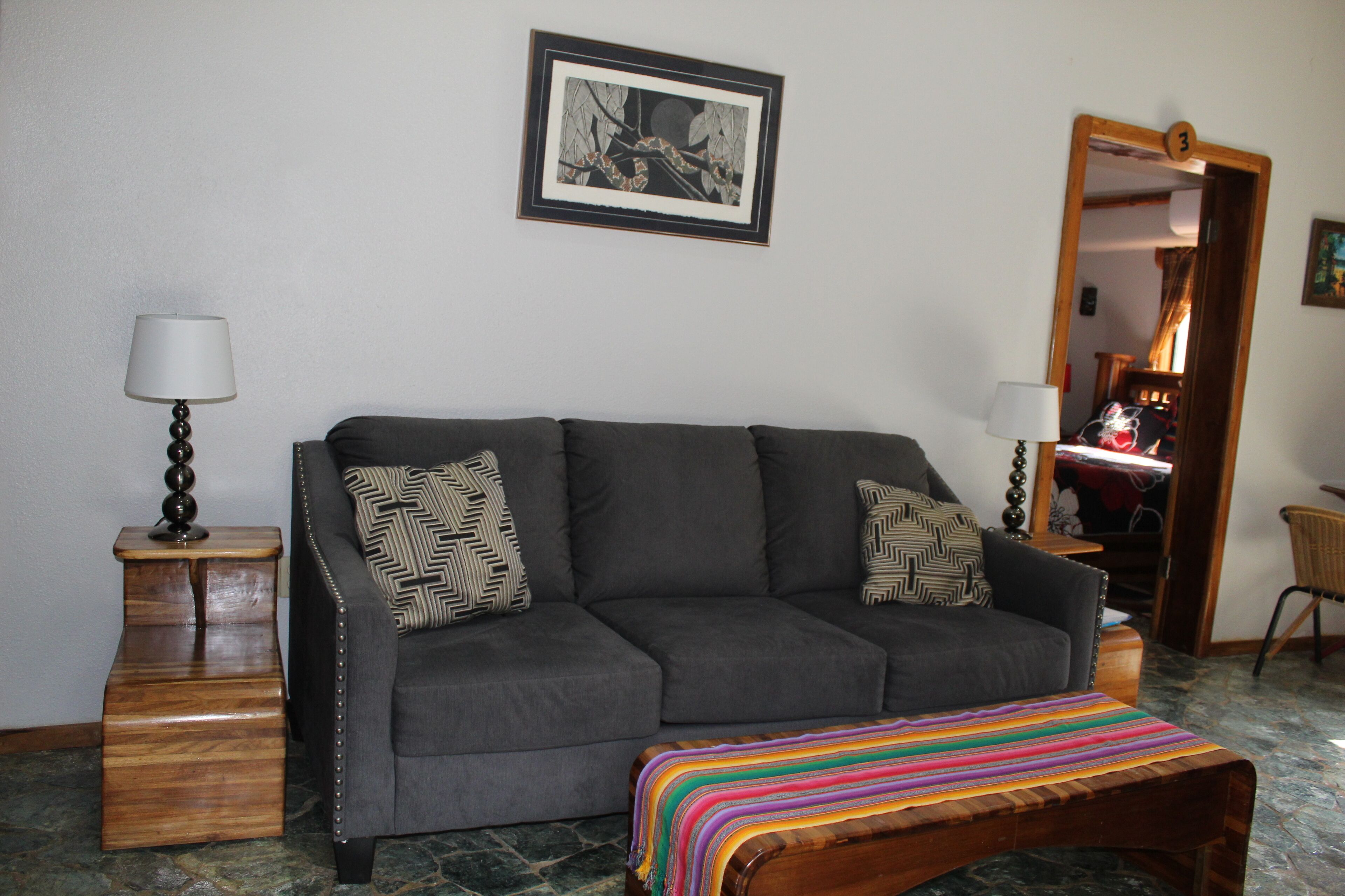 A living room with a dark grey sofa, patterned cushions, a wooden side table with a lamp, a colorful striped bench, a framed picture on the wall, and a mirror with a number on it.