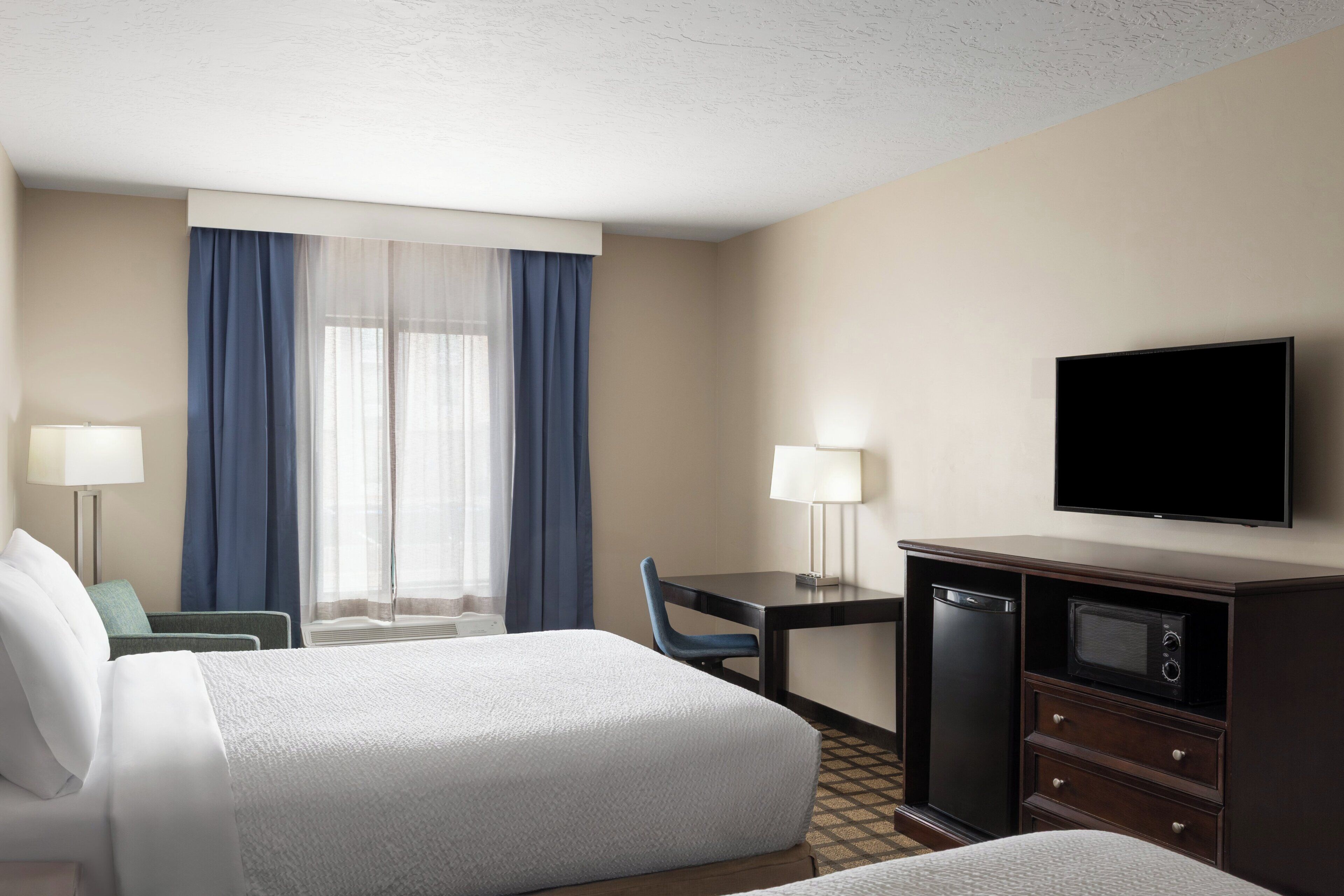 A hotel room with two beds, a desk with a television, a microwave, and a window with curtains.